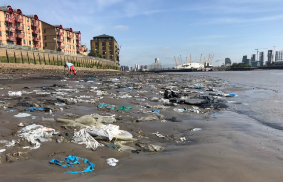 Litter and debris scattered across a muddy urban riverbank, with distant cityscape, including a domed structure, and residential buildings in the background.