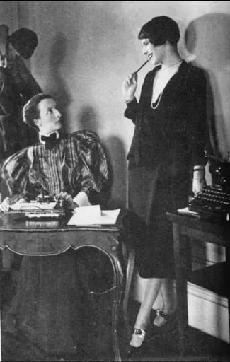 Two women in 1920s attire. One sits at a desk with papers, the other stands holding a pen, both engaged in a conversation in an office setting.