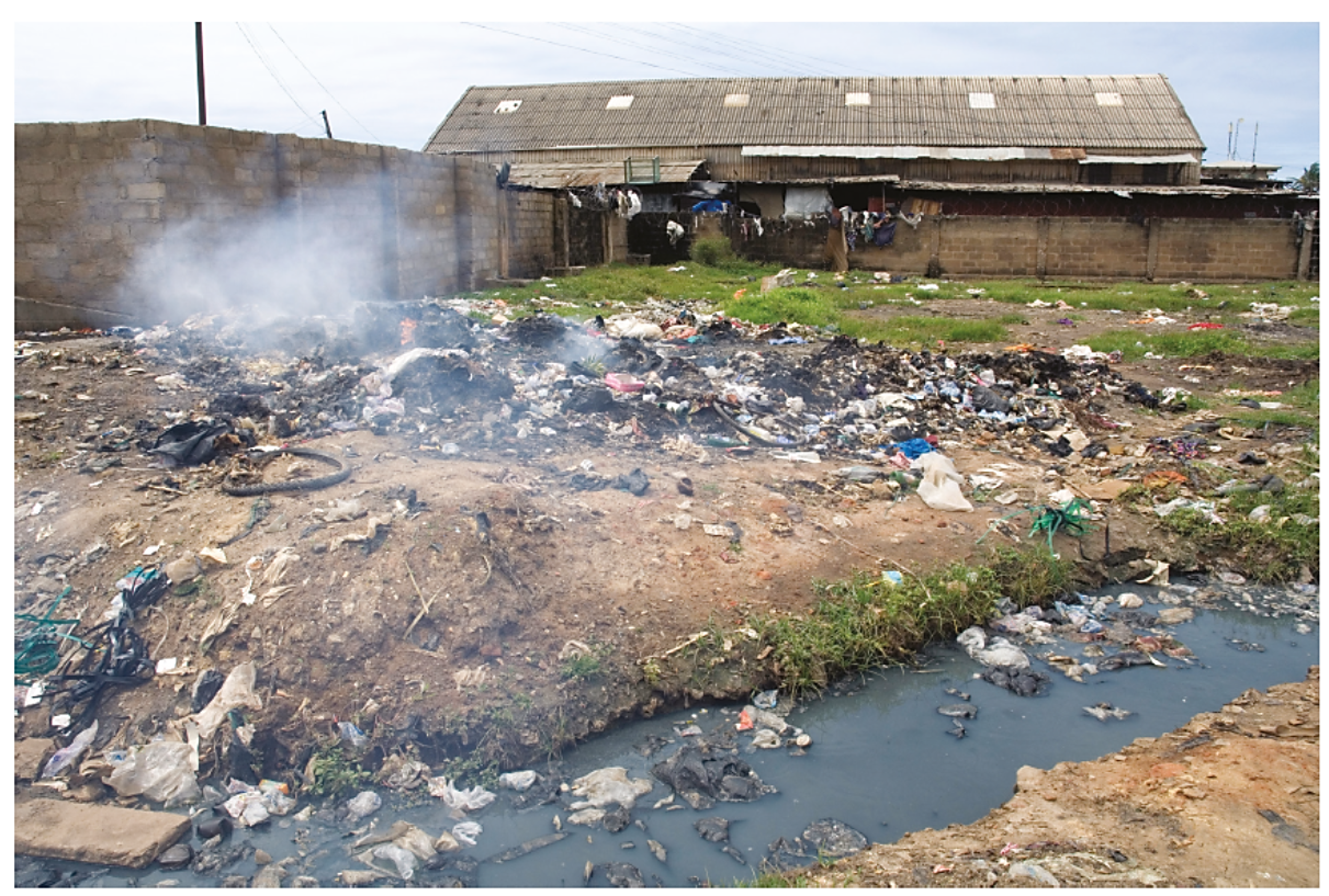 Outdoor landfill with smoking waste piles, scattered rubbish, and stagnant water near a brick building with a corrugated metal roof.