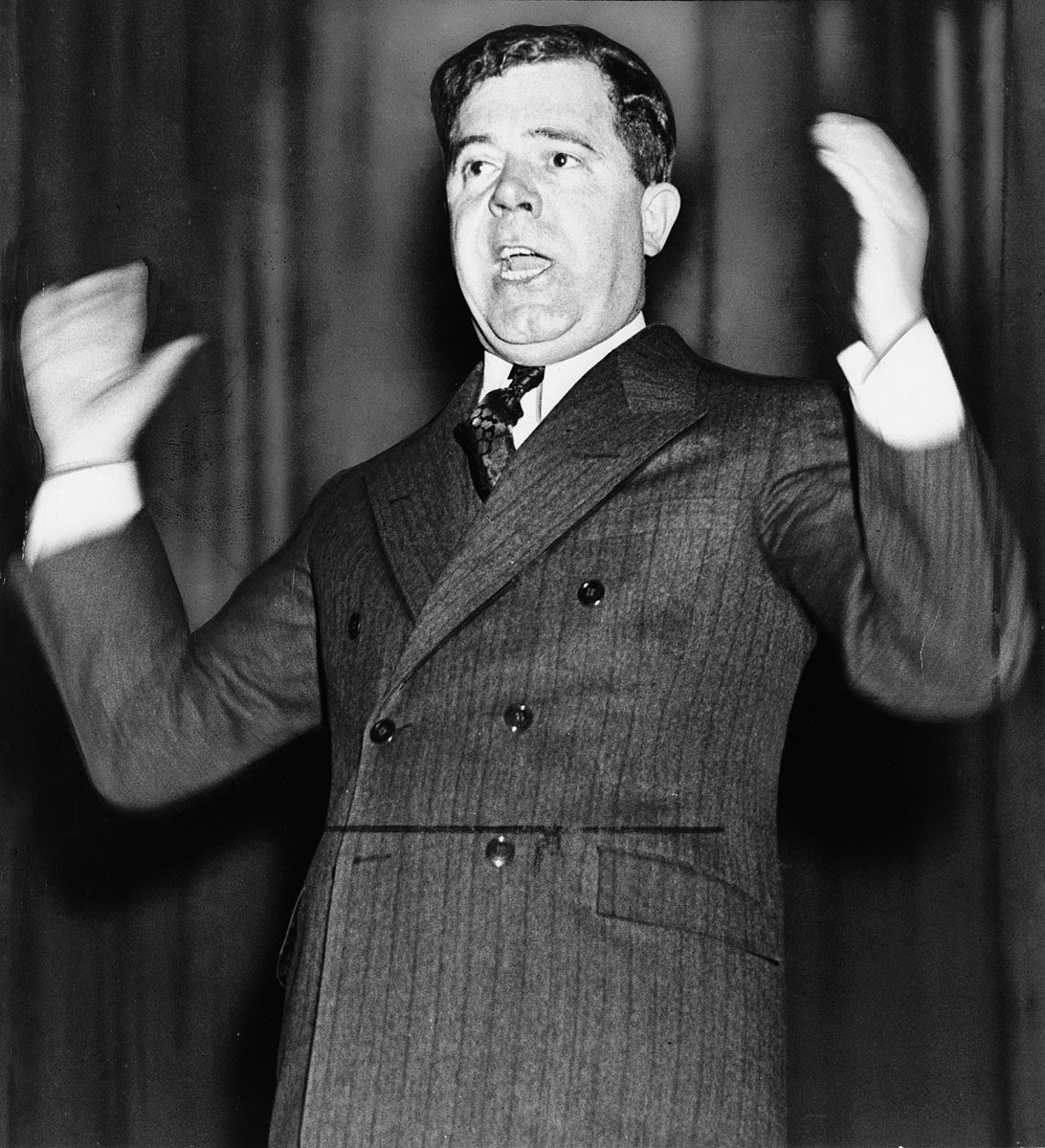 Man in a suit making a speech with expressive hand gestures, appearing engaged and energetic, set against a blurred dark background.