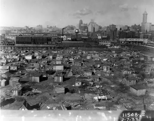 Depression-era shantytown with numerous makeshift huts, set against an urban skyline with various large buildings, showing economic hardship.