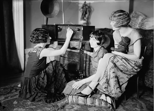Three women with bobbed hair sit around a vintage radio in a living room. One adjusts the controls, while another holds a teddy bear.
