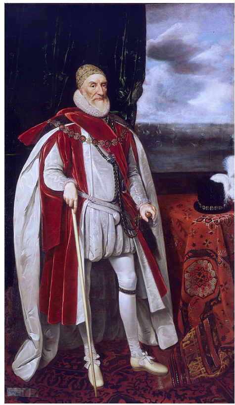 An older man in regal attire stands holding a walking stick. He wears a red and white robe, a white ruff, and ornate jewellery, beside a table with a hat.