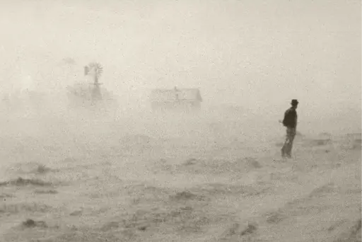A man stands alone in a barren, dust-covered landscape with a small building and a windmill faintly visible through the haze, evoking desolation.