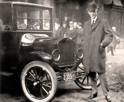Man in a suit and bowler hat stands beside a vintage car with visible licence plate. Background shows a blurred street scene with people.