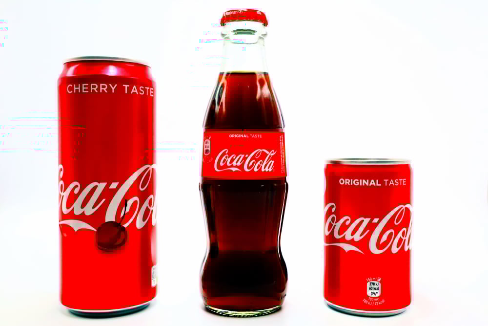 Three Coca-Cola beverages: a tall red cherry-flavoured can, a classic glass bottle, and a small red original taste can, all on a white background.