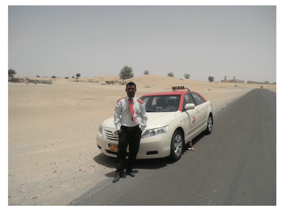 Man in white shirt and red tie stands beside a beige taxi with a red roof, parked on a desert road under a clear sky. Desert and trees in background.