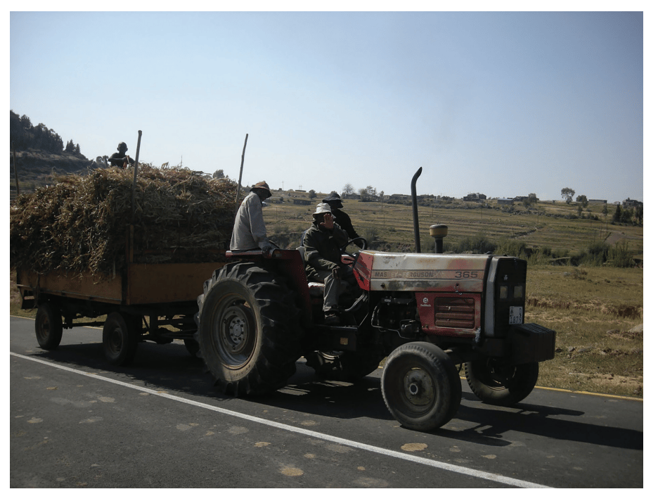 A tractor pulling a trailer filled with hay drives along a rural road, with two people seated on the tractor and fields in the background.
