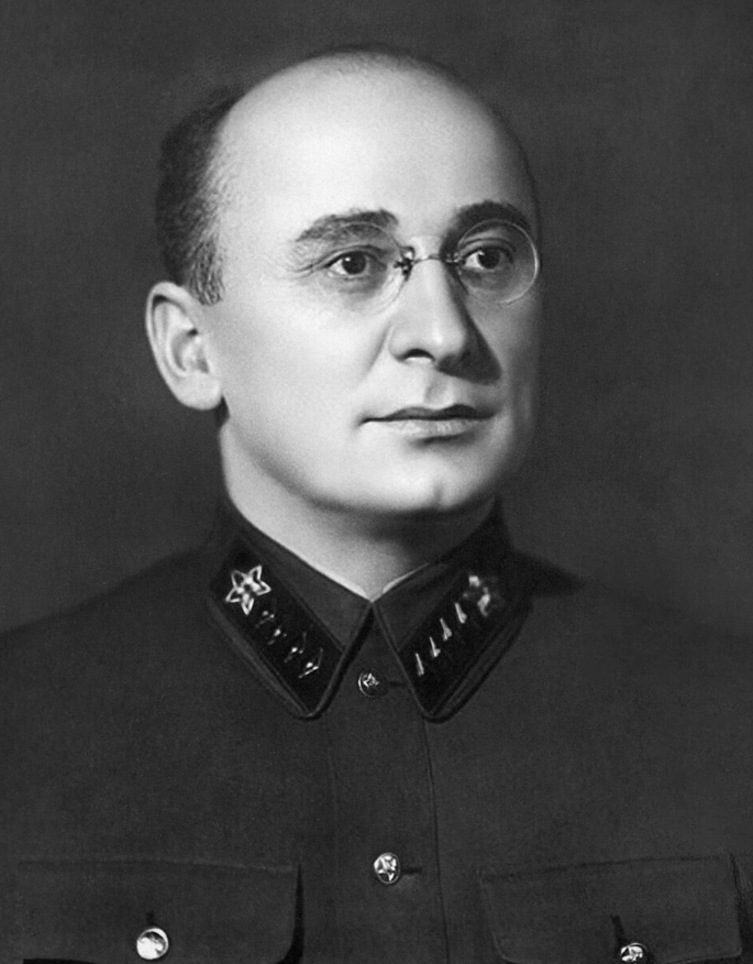 Bald man with glasses in a dark military uniform adorned with star insignia, gazing to the side against a plain backdrop.
