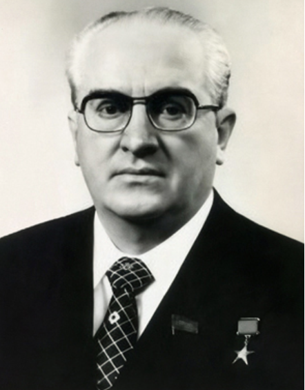 Black and white photo of an older man wearing glasses, a suit, and a patterned tie, with a medal and star badge on his lapel.