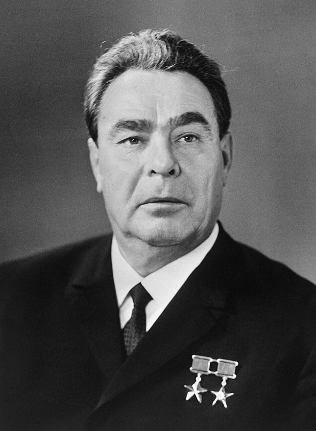 Elderly man with grey hair wearing a dark suit and tie, featuring two star medals pinned to his jacket, against a plain background.