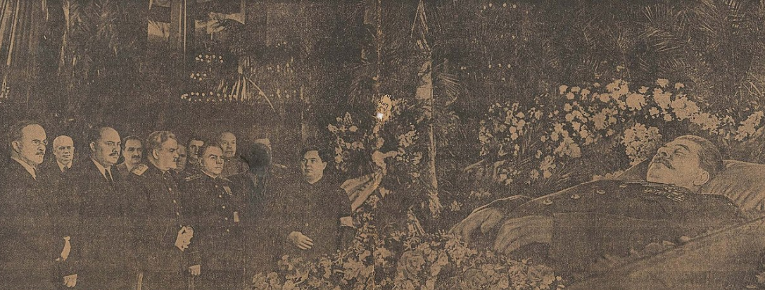 A group of people stand solemnly beside an open casket, surrounded by abundant floral arrangements in a dimly lit room.