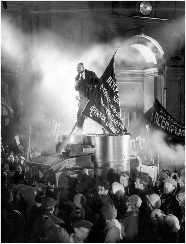 A man, depicting Lenin, giving a speech atop an armoured vehicle surrounded by a crowd, with banners and smoke, in front of the Winter's Palace in Petrograd (St Petersburg) during the October Revolution. The scene is in black and white.