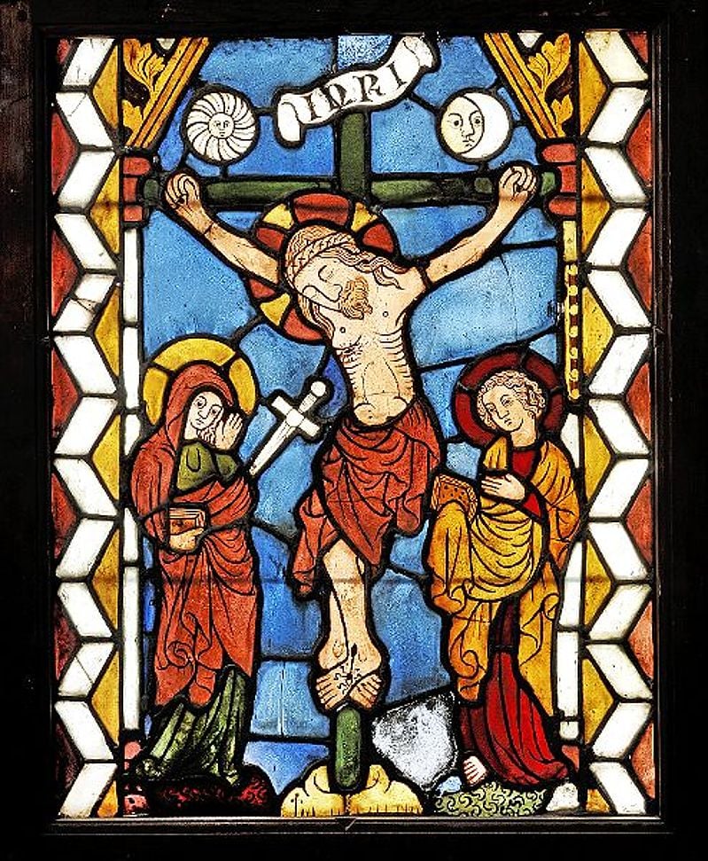 Stained glass depicting the Crucifixion, with Christ on the cross, flanked by two figures. A banner reads "INRI," set against a vibrant blue background.