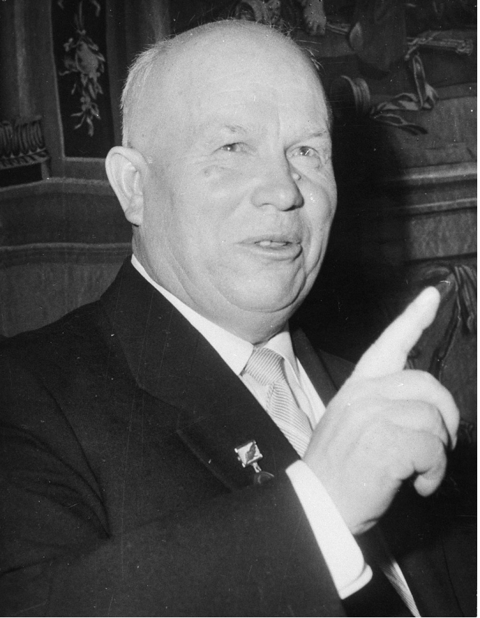 Bald man in a suit pointing upwards, smiling, with a decorative background, suggesting a formal or official setting.