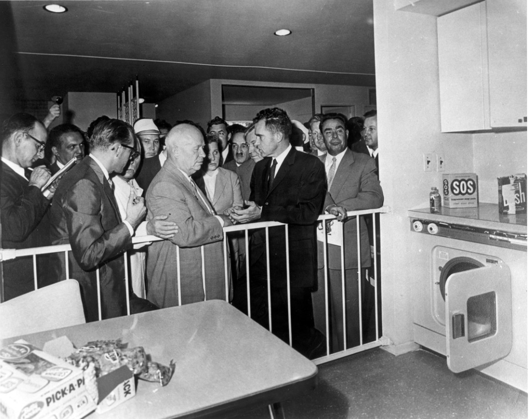 A group of men, including officials and onlookers, stand in a kitchen discussing, with household items like a washing machine and cleaning products visible.
