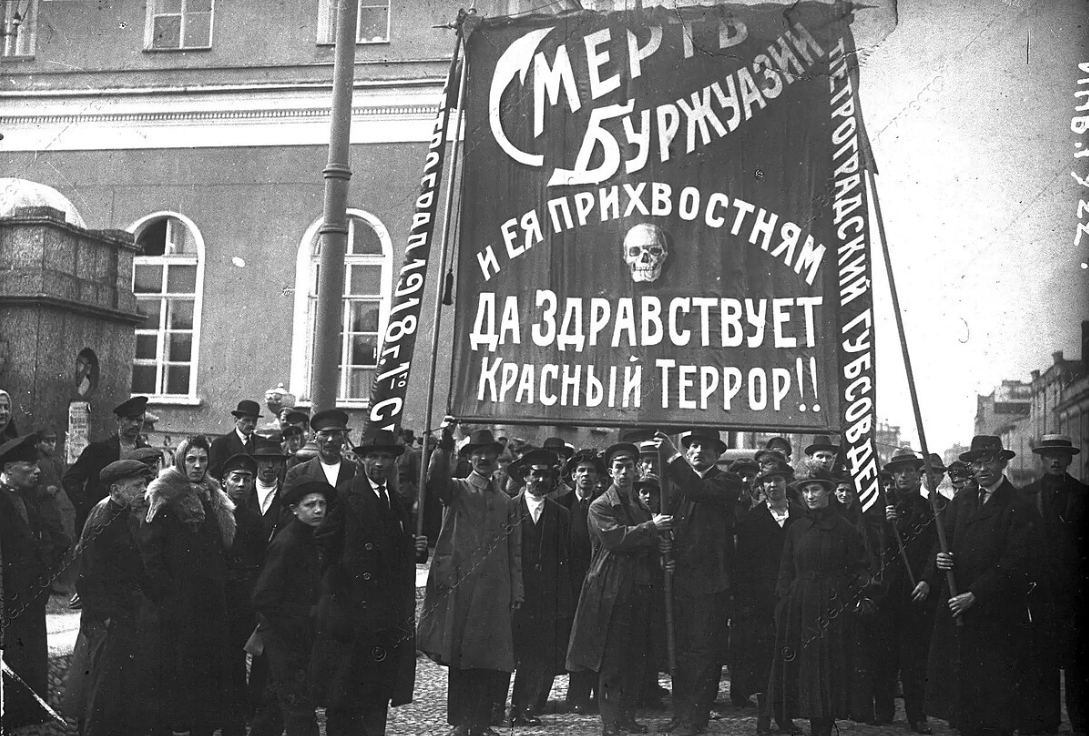 A group of people stand in a street holding a large banner with Russian text and a skull image, in front of a building with arched windows.