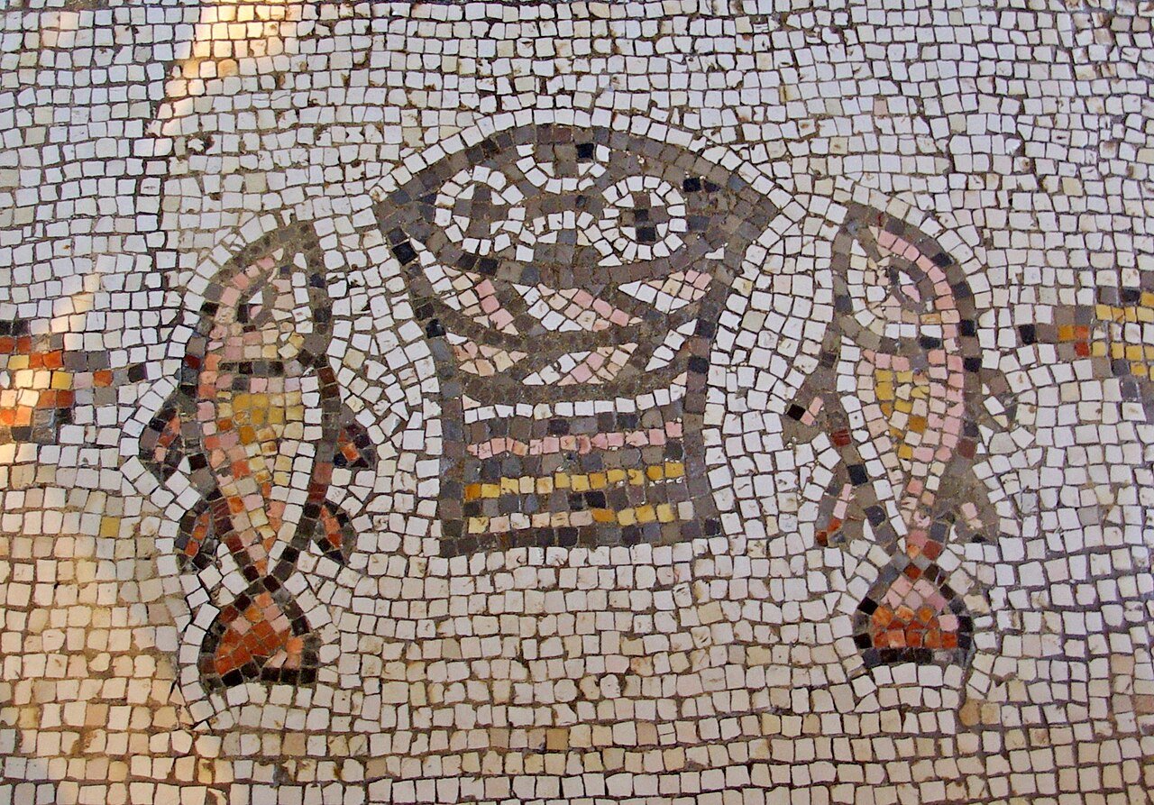 Ancient mosaic depicting two fish and a basket with loaves, crafted from small, coloured tiles on a cream background.