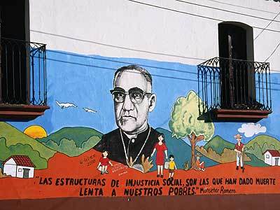 Mural on a building wall featuring a portrait of a man in a clerical collar, surrounded by a village scene with children, trees, and mountains.