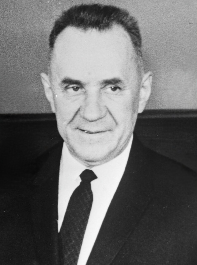 Older man in a suit and tie, smiling slightly. He has short dark hair and a light complexion. The background is plain and blurred.