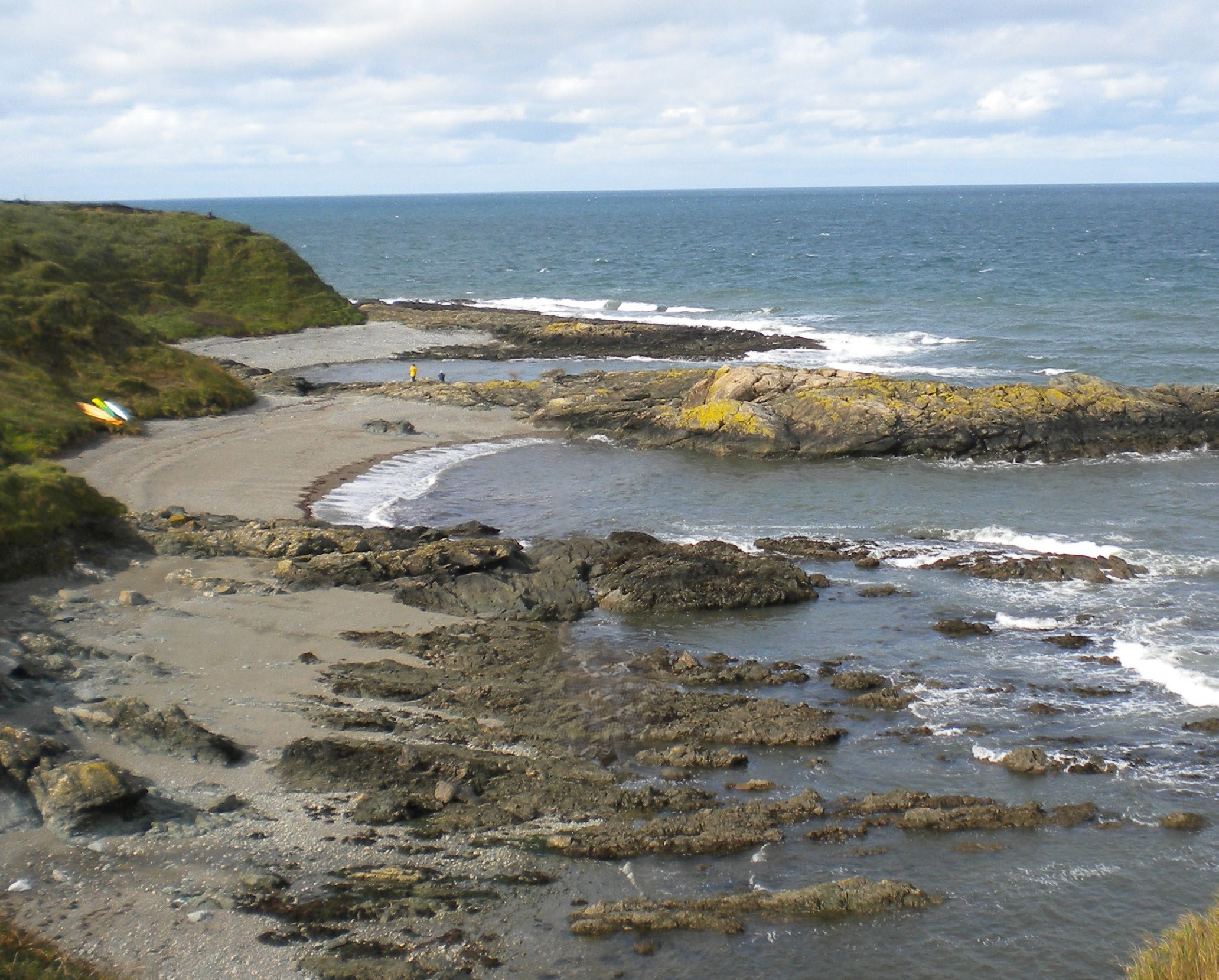 Rocky coastal landscape with small sandy beaches, sea, and grassy cliffs under a cloudy sky. A kayak is visible on the left shore.