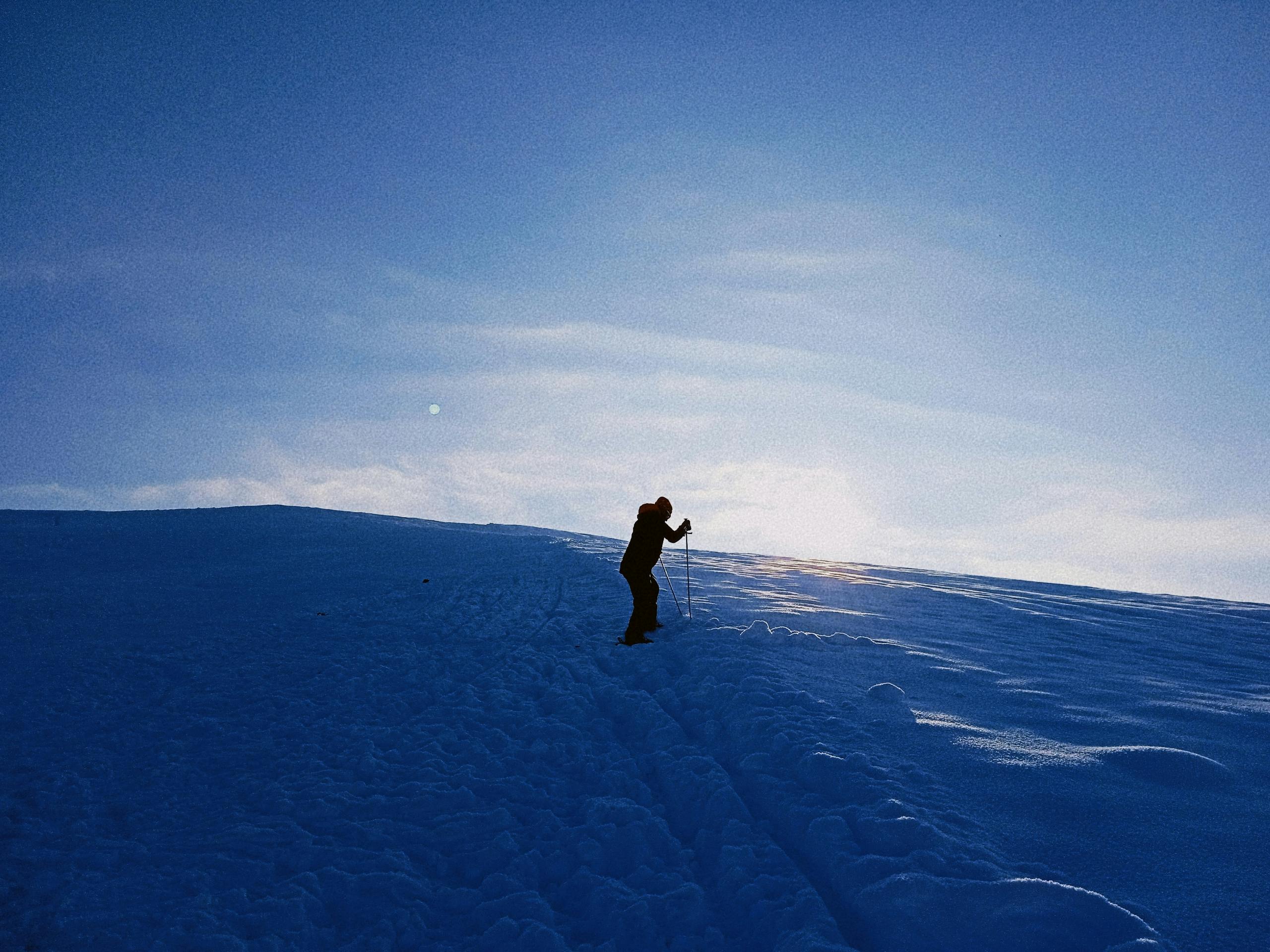 Silhouetted person skiing uphill on a snowy landscape at sunrise, with a clear blue sky and bright sunlight casting long shadows on the snow.