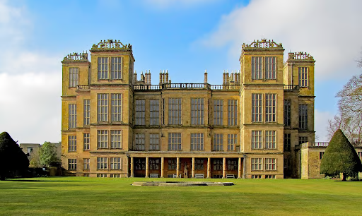 Historic manor house with a symmetrical facade, large windows, and ornate chimneys, set against a clear sky, surrounded by manicured lawns.