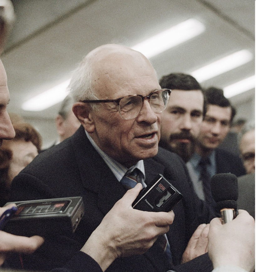 Elderly man with glasses speaking to a group of reporters holding microphones and tape recorders in a crowded indoor setting.