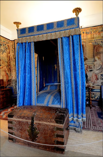 Antique four-poster bed with vibrant blue drapes, ornate wooden trunk at the foot, surrounded by detailed tapestry wall hangings in a historic room.