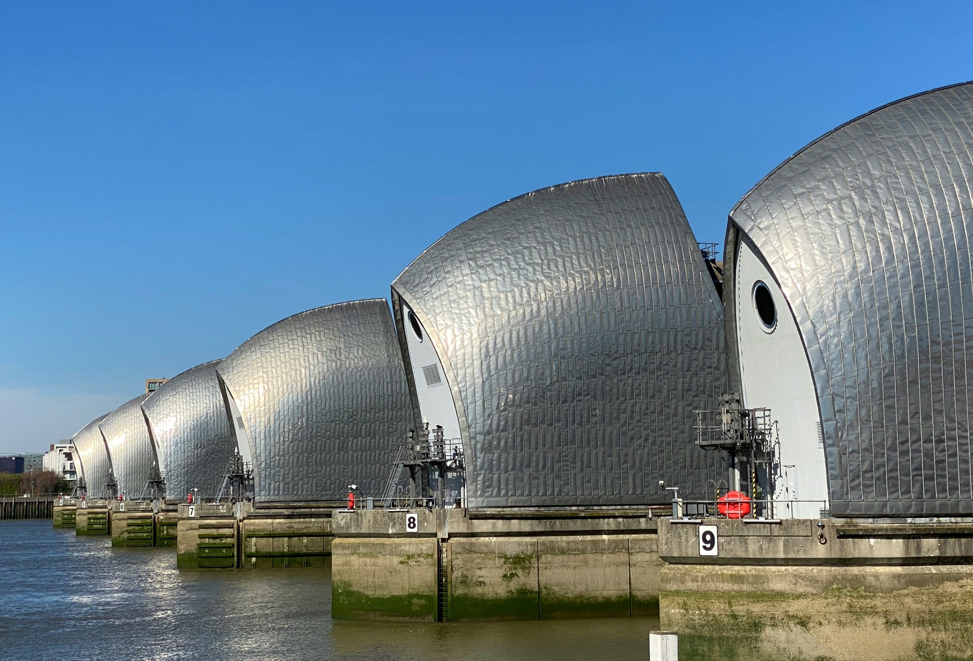 Silver arched barriers of the Thames Barrier on a sunny day, with blue sky above and river water below, featuring numbered sections.