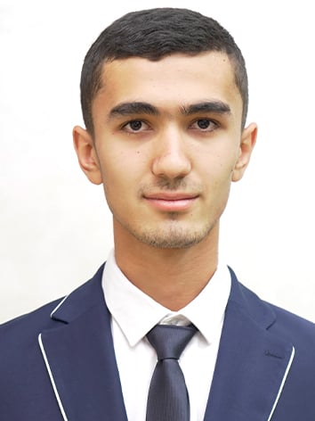 Young man with short dark hair wearing a dark suit and tie, with a white shirt and light piping on the jacket's lapels, against a plain background.