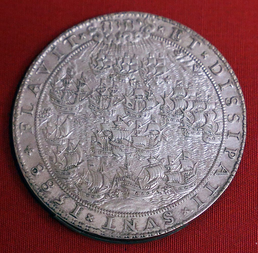 Silver coin displaying a detailed engraving of a naval battle with numerous sailing ships, surrounded by an inscribed border, set against a red background.