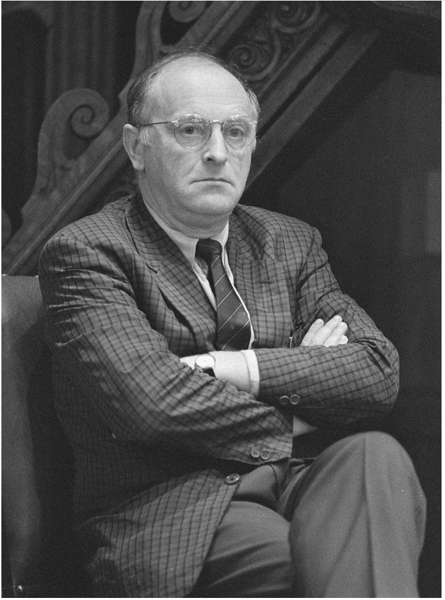Elderly man with glasses sits with arms crossed, wearing a checkered suit and tie. He appears serious and is in front of ornate wooden railing.