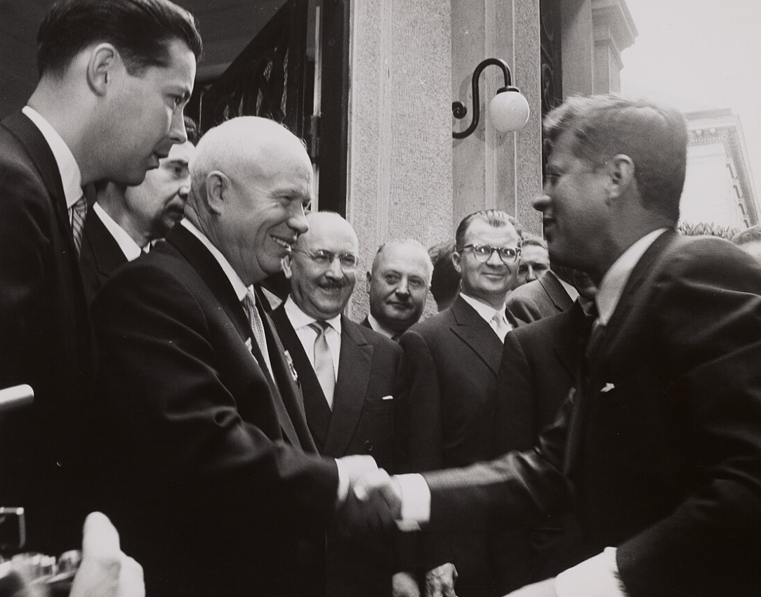 A group of men in suits gather near a building, while two prominent figures in the foreground exchange a handshake and smile.