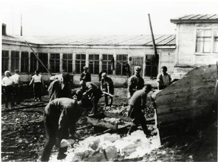 Historic black and white photo: group of people engaged in manual labour, possibly construction, in front of a large building with multiple windows.