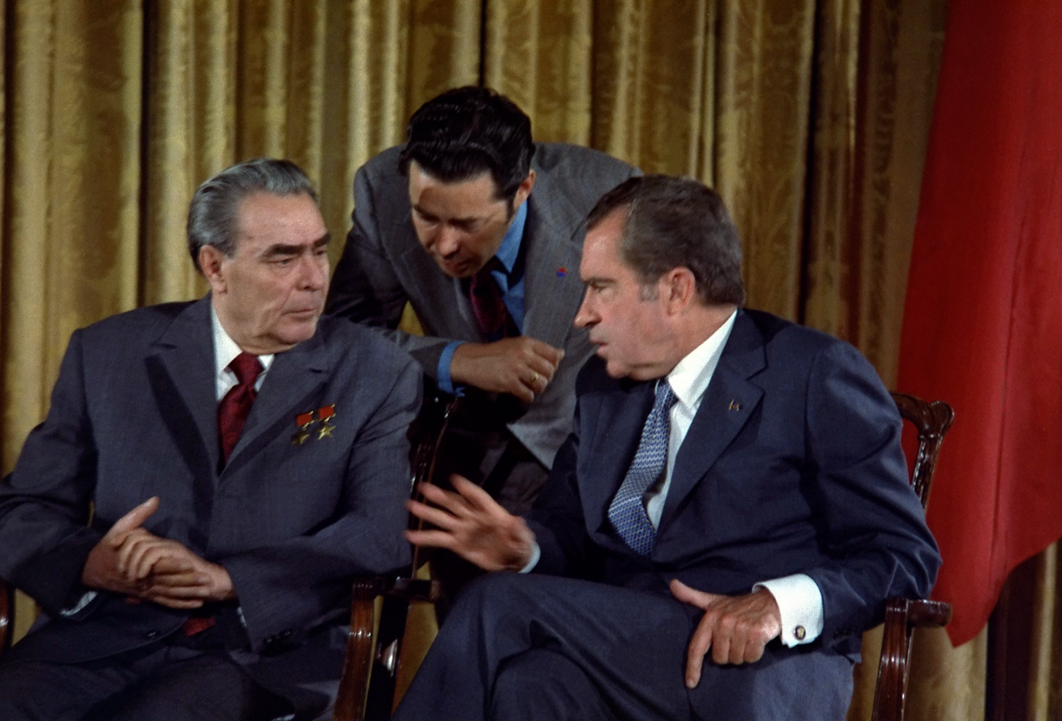 Two men in suits are conversing, seated in chairs with a third man leaning in to listen. A gold curtain and red flag are in the background.