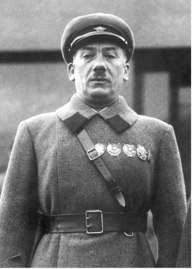 Uniformed man with military cap and several medals, standing outdoors. He wears a belted grey coat with contrasting collar. Background is blurred.