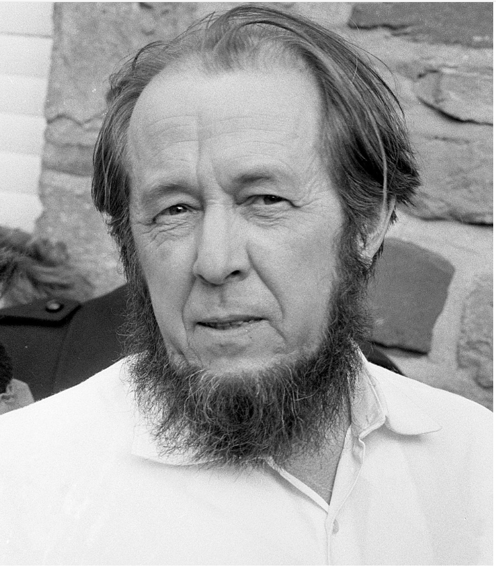Older man with a long beard and receding hairline, wearing a white shirt outdoors. Stone wall in the background, conveying a sombre expression.