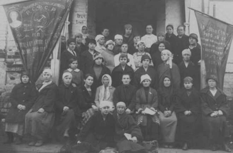 Group photo of women in early 20th century attire, gathered outside a building with two banners featuring Cyrillic text on either side.