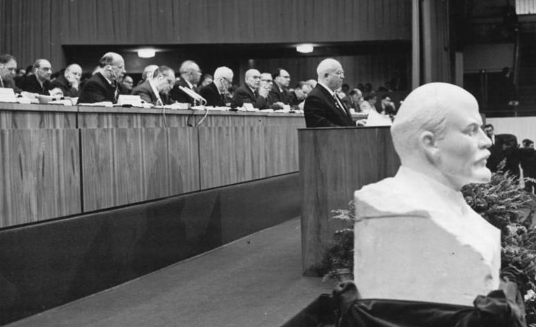 A conference hall with a row of seated men in suits, a speaker at a podium, and a prominent bust of Lenin in the foreground.
