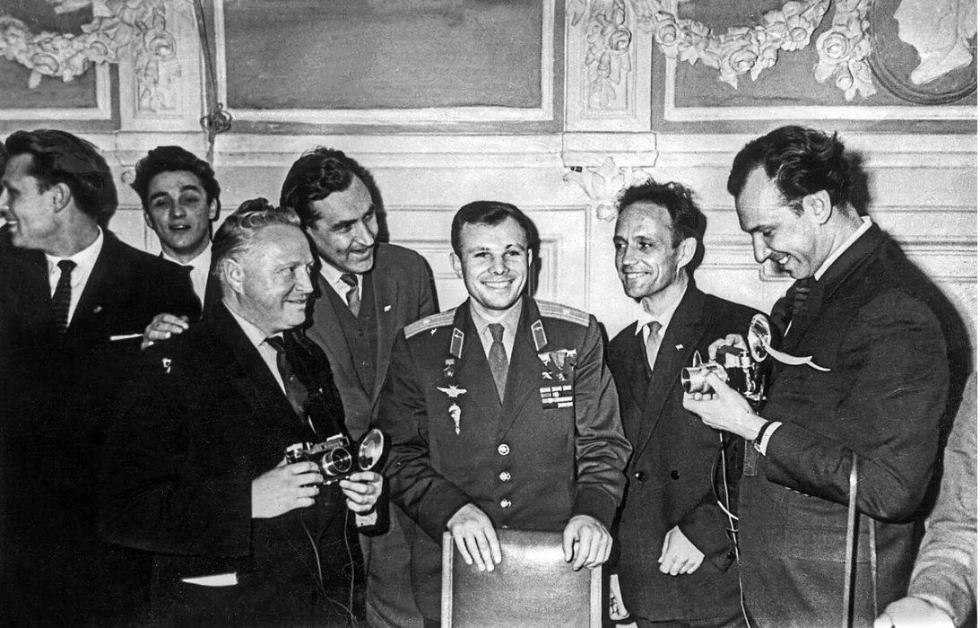 Six men in formal attire with one in a decorated military uniform stand together, smiling, in an ornately decorated room; two hold cameras.