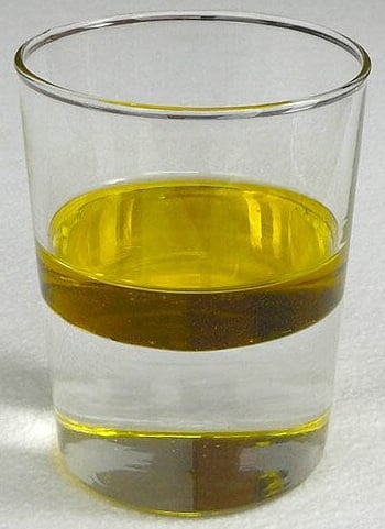 A clear glass filled with water and a layer of yellow oil floating on top, demonstrating liquid density differences on a light surface.