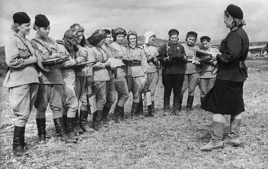 A group of women in military uniforms and flight gear stand in formation outdoors, holding notebooks and listening to a woman addressing them.