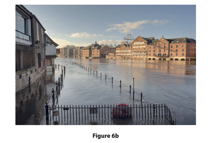 Flooded riverside path with partially submerged railings and buildings, including a visible "Warehouse" sign, under a clear sky.
