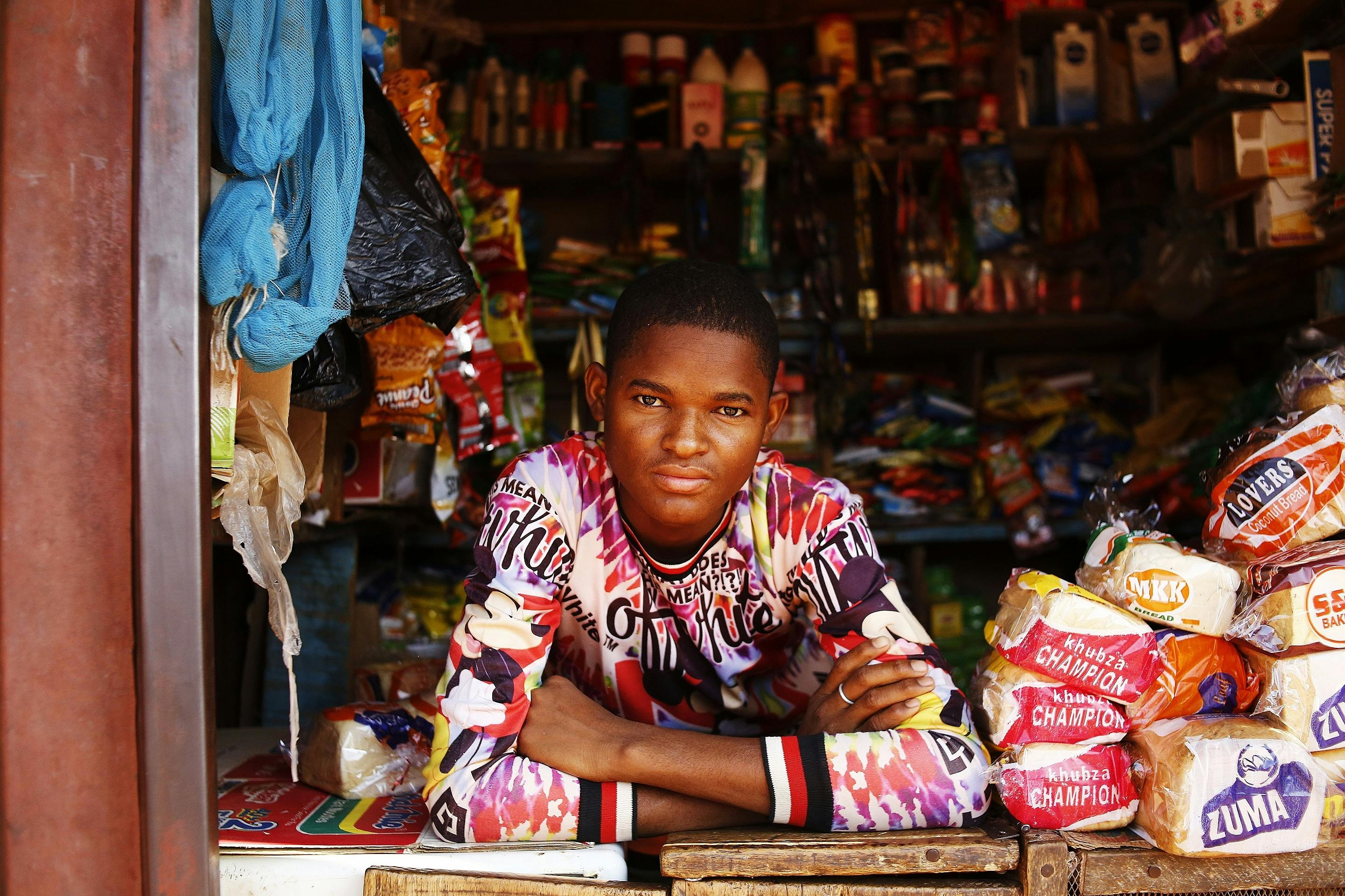 Young man in a colourful shirt leans on a wooden counter in a cluttered shop with various packaged goods and household items displayed around him.
