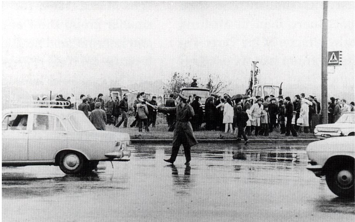 A group of people gathered by the roadside near parked cars and a tractor, with a coat-wearing person directing traffic on a wet street.