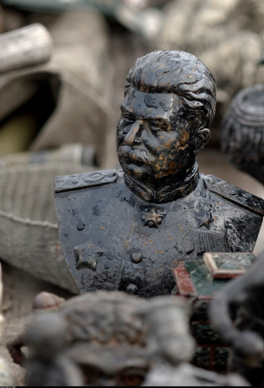 Weathered bust of a military figure with a moustache, wearing decorated uniform, amidst various worn objects and blurred background.