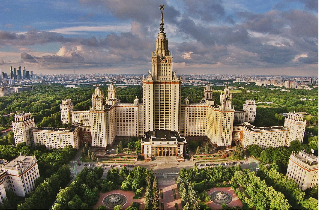 Aerial view of a grand neoclassical skyscraper surrounded by greenery and cityscape. The central tower is tall and ornate, topped with a spire bearing a Soviet-style star emblem. The main structure has symmetrical wings extending from both sides, each adorned with clock towers and decorative detailing. The building is cream-colored with red and gold accents, and a large courtyard and gardens stretch out in front. In the background, a dense forest and modern city skyline are visible under a dramatic, partly cloudy sky.