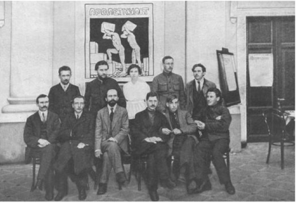 Group of eleven men and one woman, seated and standing, in front of a poster with industrial imagery in a hall with columns and an easel.