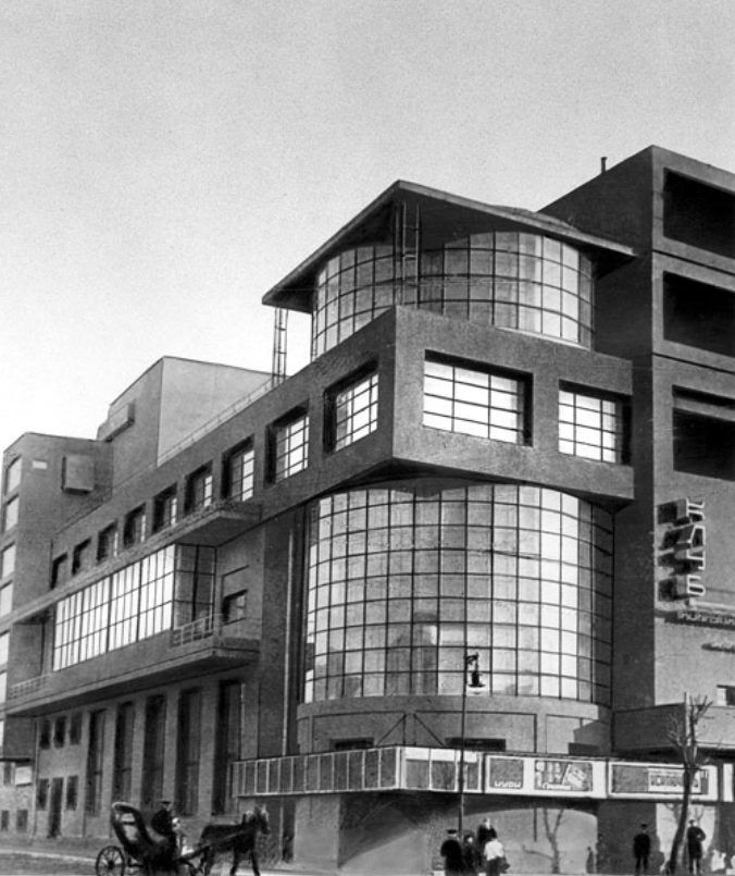 Historic black and white photo of a modernist building with large glass windows, curved façades, and a horse-drawn carriage in the foreground.